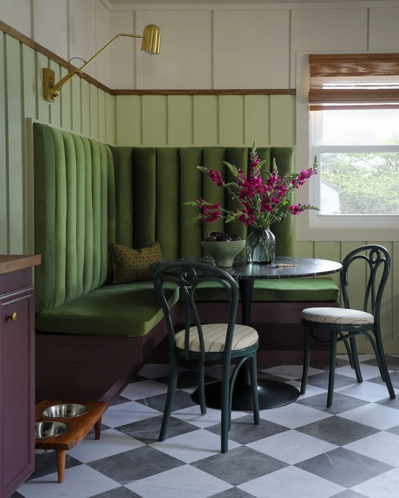 A breakfast nook with a green velvet banquette with high backrest, round black table, two black chairs, floral centerpiece, checkered floor, and a window with a bamboo shade.