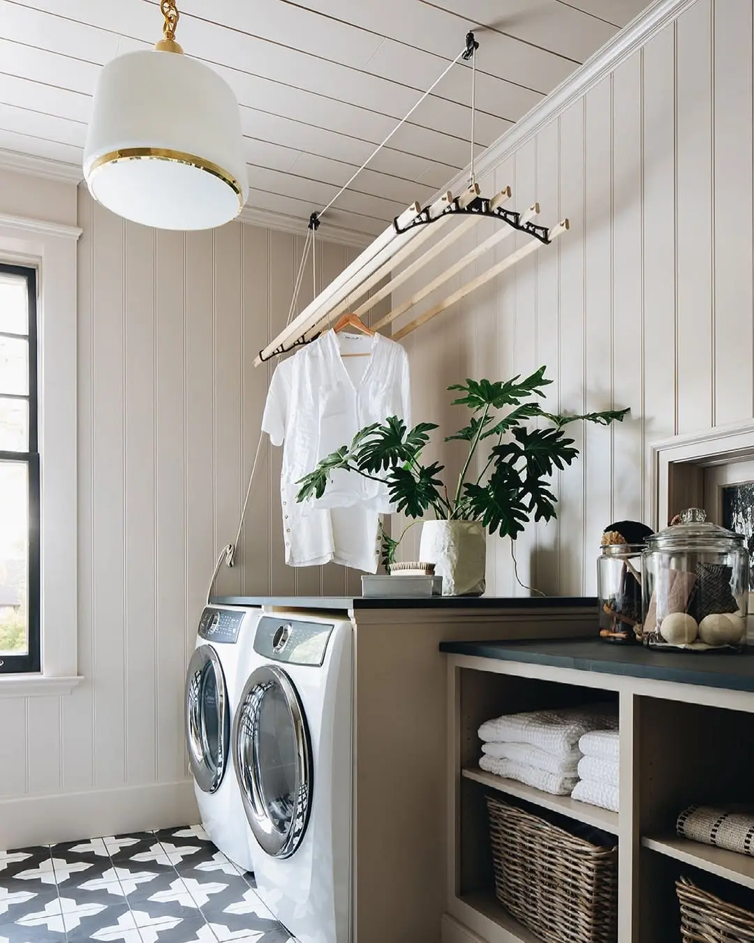 A modern laundry room with a front-loading washer and dryer, a hanging rack with clothes, a plant on the counter, and shelves with neatly folded towels and baskets, showcasing smart laundry storage ideas.