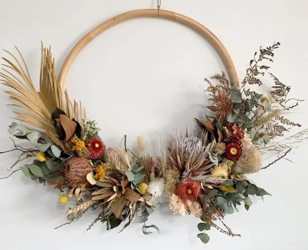 A large wooden hoop decorated with an arrangement of dried flowers, leaves, and grasses in earthy winter tones, hanging against a plain white wall.