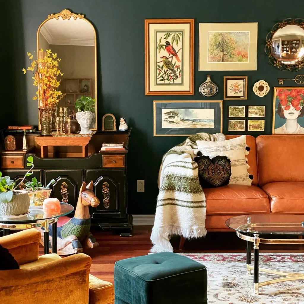 Eclectic living room with green walls, brown leather sofa, gallery wall art, gold mirror, vintage decor, plants, and a playful llama figurine. Cozy blanket and pillows complete this inviting, eclectic living room vibe.