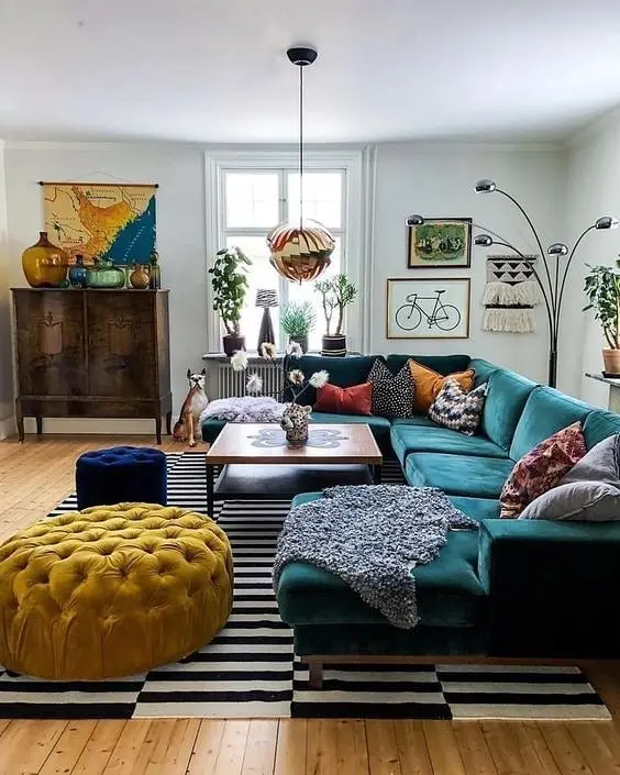 A colorful, eclectic living room with a teal sectional sofa, yellow tufted ottoman, patterned pillows, striped rug, houseplants, and vibrant wall art and decor.