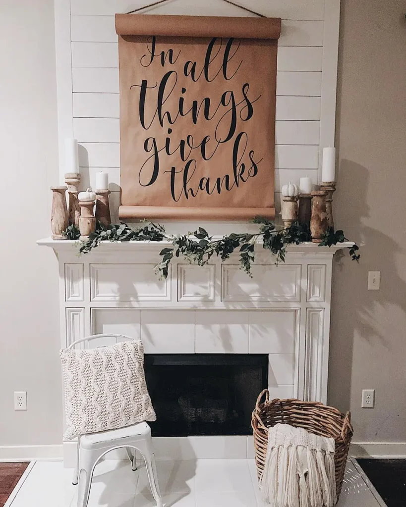 A white fireplace mantel decorated with candles, greenery, earthy fall tones, a sign reading "In all things give thanks," a white chair with a textured pillow, and a wicker basket with a blanket.