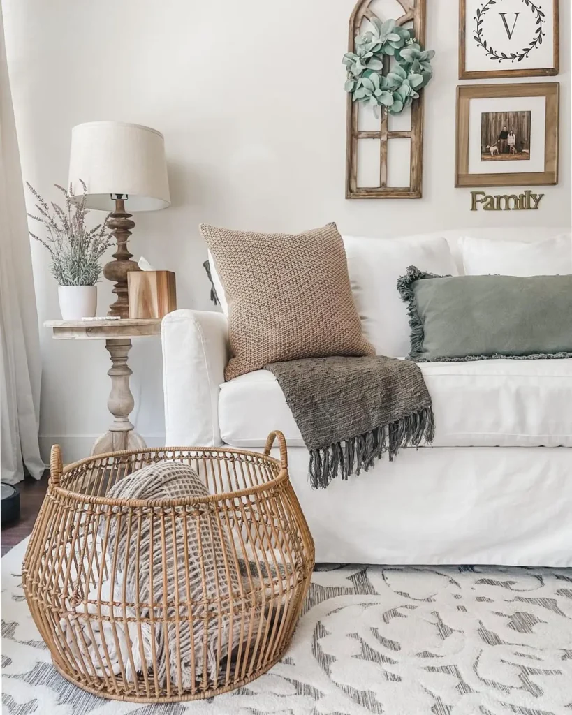 A neutral-toned living room with a white sofa, green and beige cushions, a side table with a lamp, wall decor, and a wicker basket with a blanket on a patterned rug, great example of how to style a farmhouse living room for fall with earthy tones.