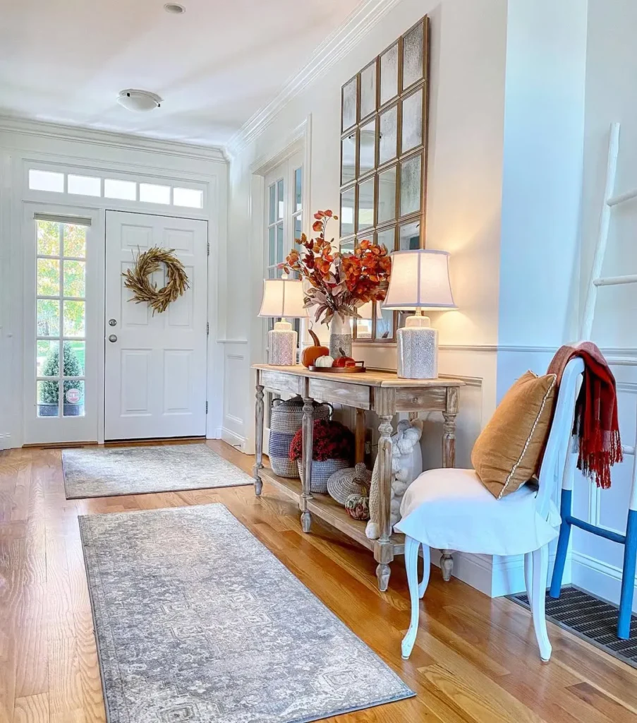 Bright entryway with double doors, a console table decorated with lamps and autumn accents, a mirror above, a white chair with a pillow and throw, and neutral rugs on a wooden floor.