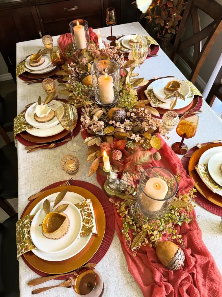 A dining table set for six with autumn-themed decorations, candles, gold accents, amber glassware, and a centerpiece of leaves, pinecones, and flowers on a rust-colored runner.