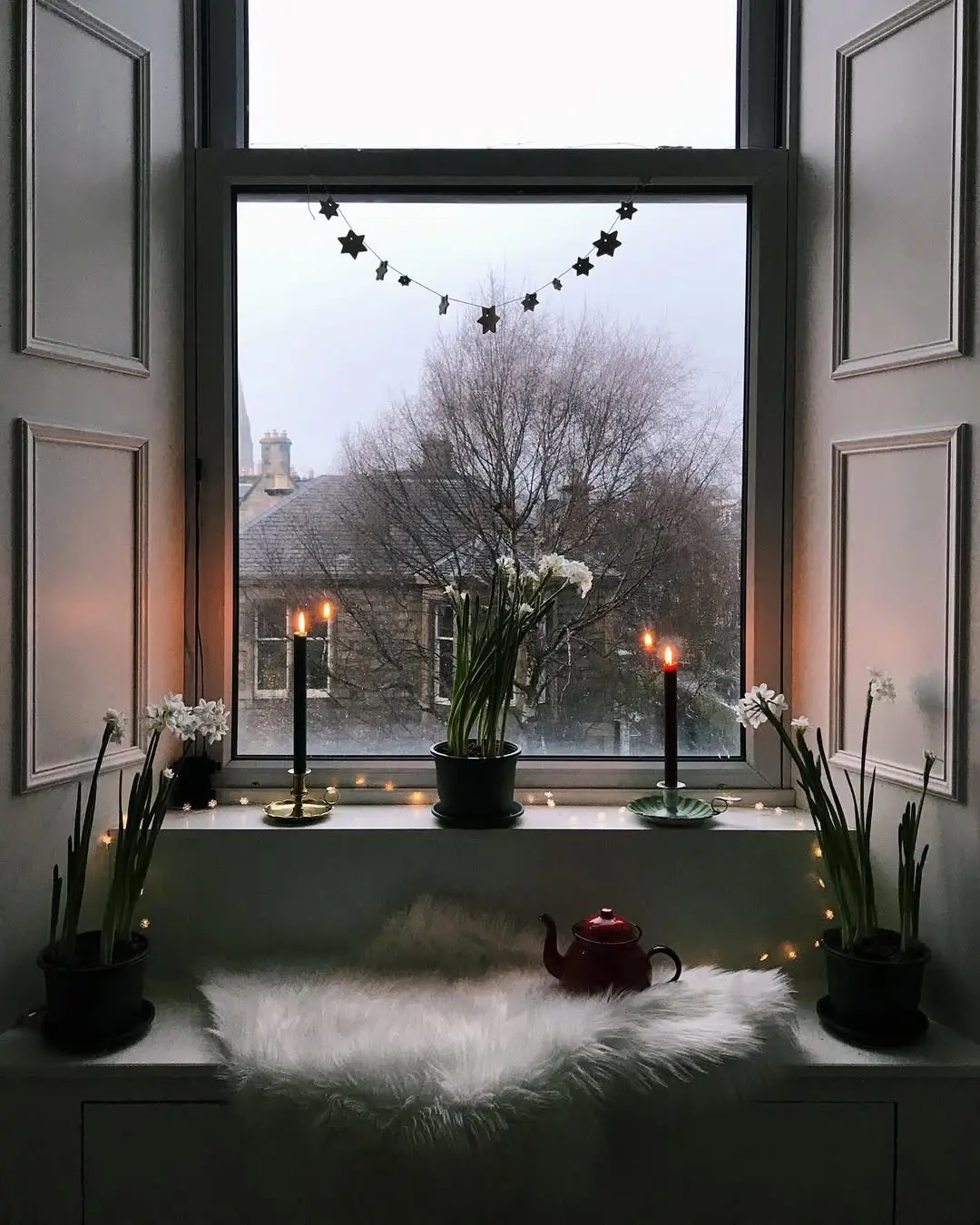 A cozy window nook with potted plants, two lit candles, string lights, a red teapot, and a furry white mat overlooking bare trees, perfect inspiration for how to transition your home decor from fall to winter on a cloudy day.
