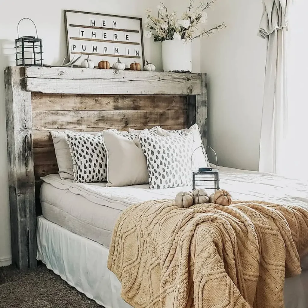 A cozy farmhouse fall bedroom with a wooden headboard, white and patterned pillows, a beige knit blanket, small pumpkins, and a "HEY THERE PUMPKIN" sign on the shelf above the bed.
