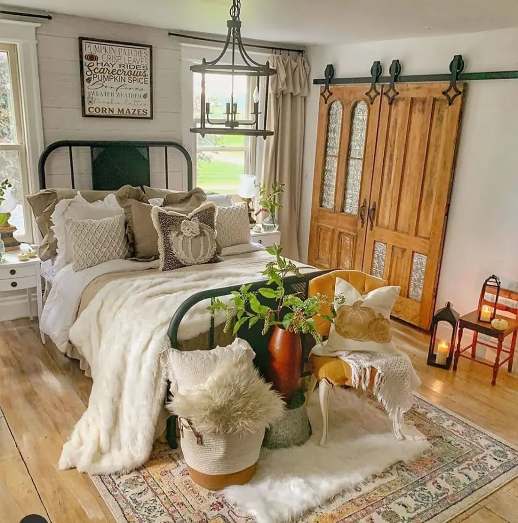 Cozy farmhouse fall bedroom with a metal-frame bed, plush pillows, throw blankets, wooden sliding barn doors, a chandelier, potted plants, and decorative lanterns on a vintage rug.