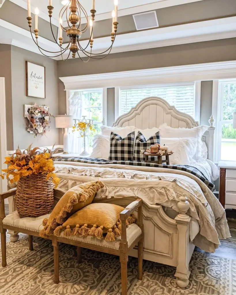 A neatly made bed with a plaid pillow stands in a bright farmhouse fall bedroom featuring a chandelier, decorative pillows, a bench with cushions, and a basket of autumn leaves at the foot of the bed.