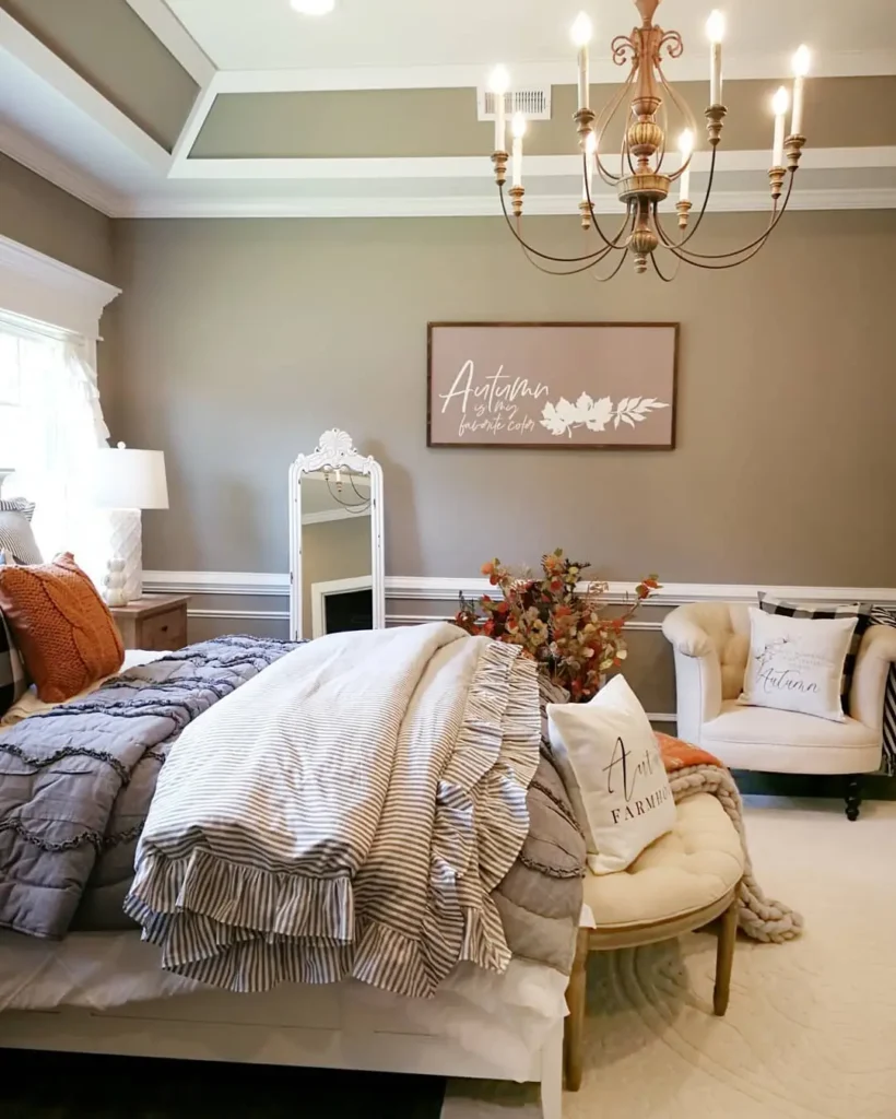 A neatly made bed with gray bedding, farmhouse fall pillows, and a throw sits in a cozy bedroom with neutral walls styled with seasonal wall art, a chandelier, an armchair, and autumn decor.