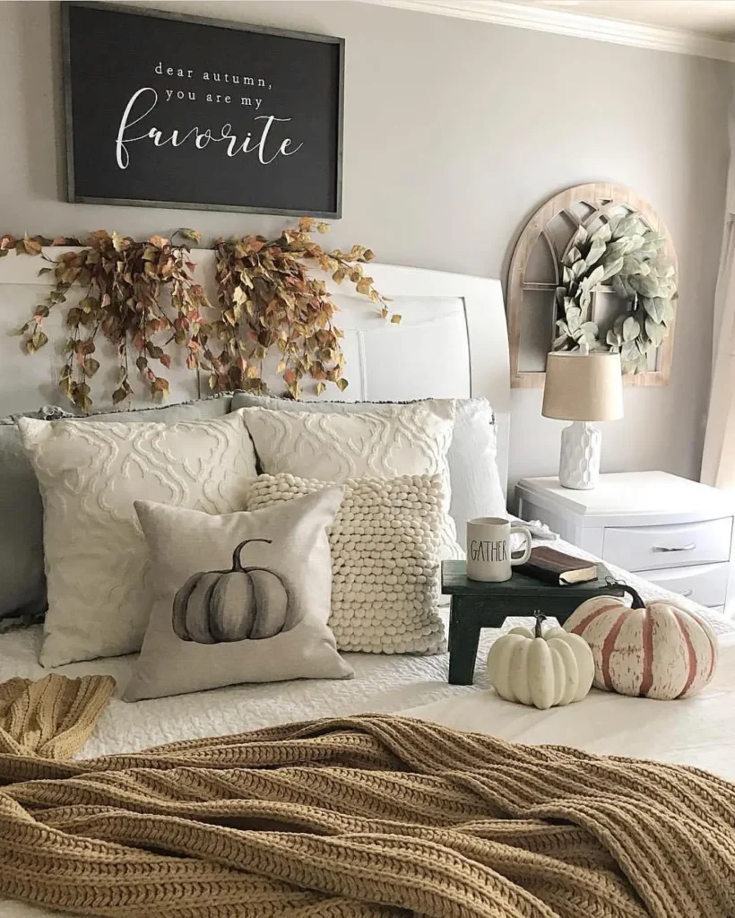 A cozy farmhouse fall bedroom decorated for autumn with white bedding, fall-themed throw pillows, faux pumpkins, and leafy garlands. A sign above the bed reads, "dear autumn, you are my favorite.