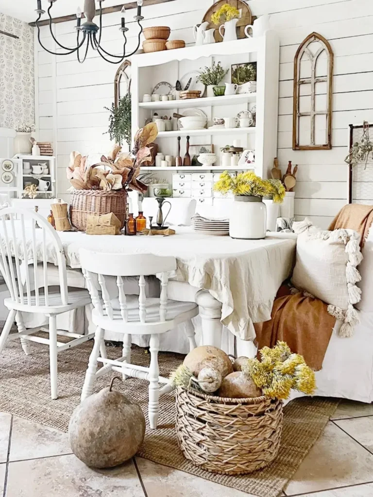 A cozy farmhouse fall dining area with a white table and chairs, neutral decor, rustic shelves, dried flowers, and woven baskets filled with gourds and yellow blooms.