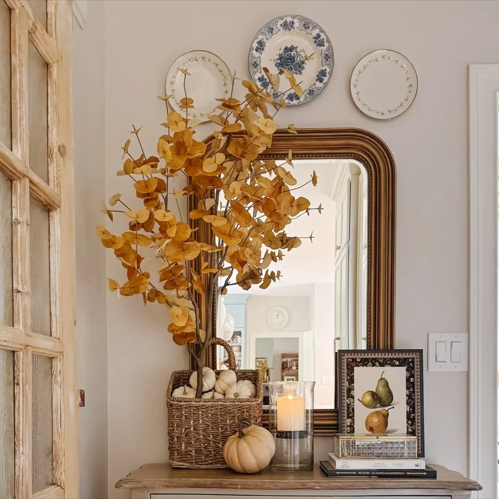 A farmhouse fall entryway decor features a console table with a basket of yellow leaves, a small pumpkin, books, a candle, and framed art, all set below a mirror and three decorative plates on the wall.
