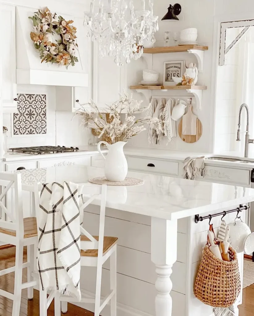 A white farmhouse kitchen decorated for fall featuring a rustic wreath, crystal chandelier, ceramic pitcher with autumn foliage, and a plaid dish towel draped over a chair for a cozy seasonal touch.