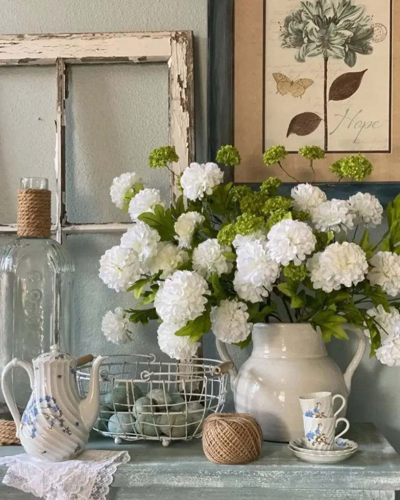 A white ceramic pitcher with white flowers, a teapot, teacup, farmhouse wire basket filled with blue eggs, and a glass bottle on a blue table; framed art and a rustic window frame in the background.