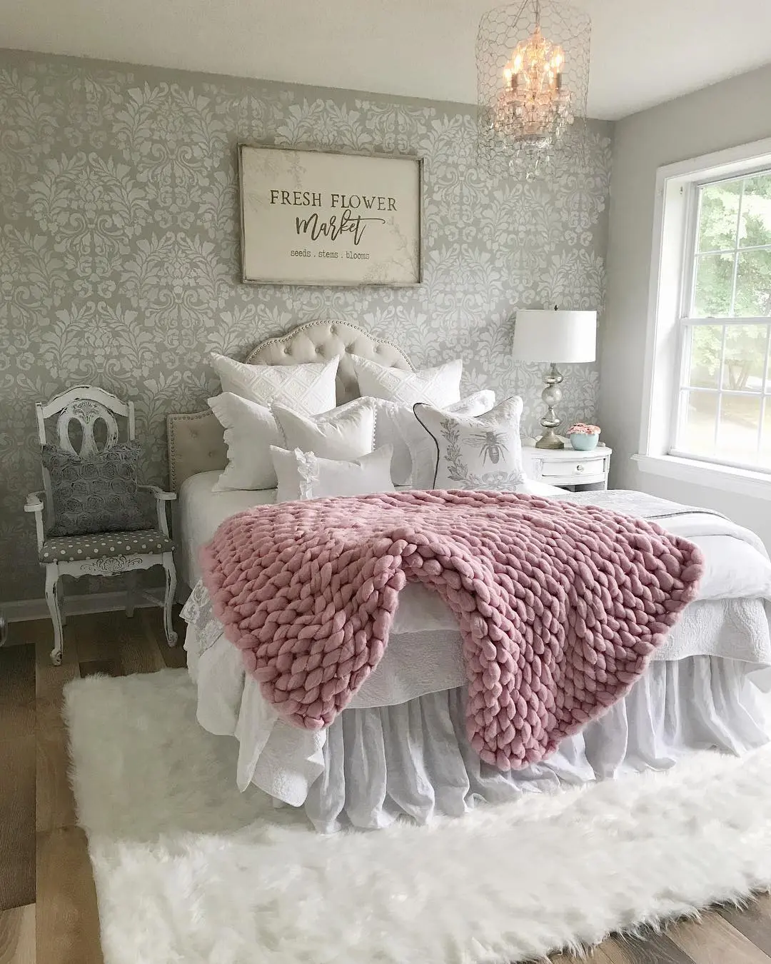A cozy bedroom with a white bed over a faux fur rug, pink chunky knit blanket, patterned accent wall, chandelier, white chair, and large window letting in natural light.