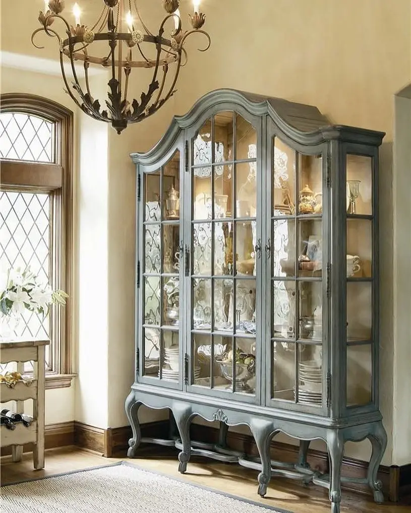 A blue-gray vintage glass-front china cabinet filled with dishes and decorative items stands against a beige wall beneath a wrought-iron chandelier, a color palette inspired by French Country decor.