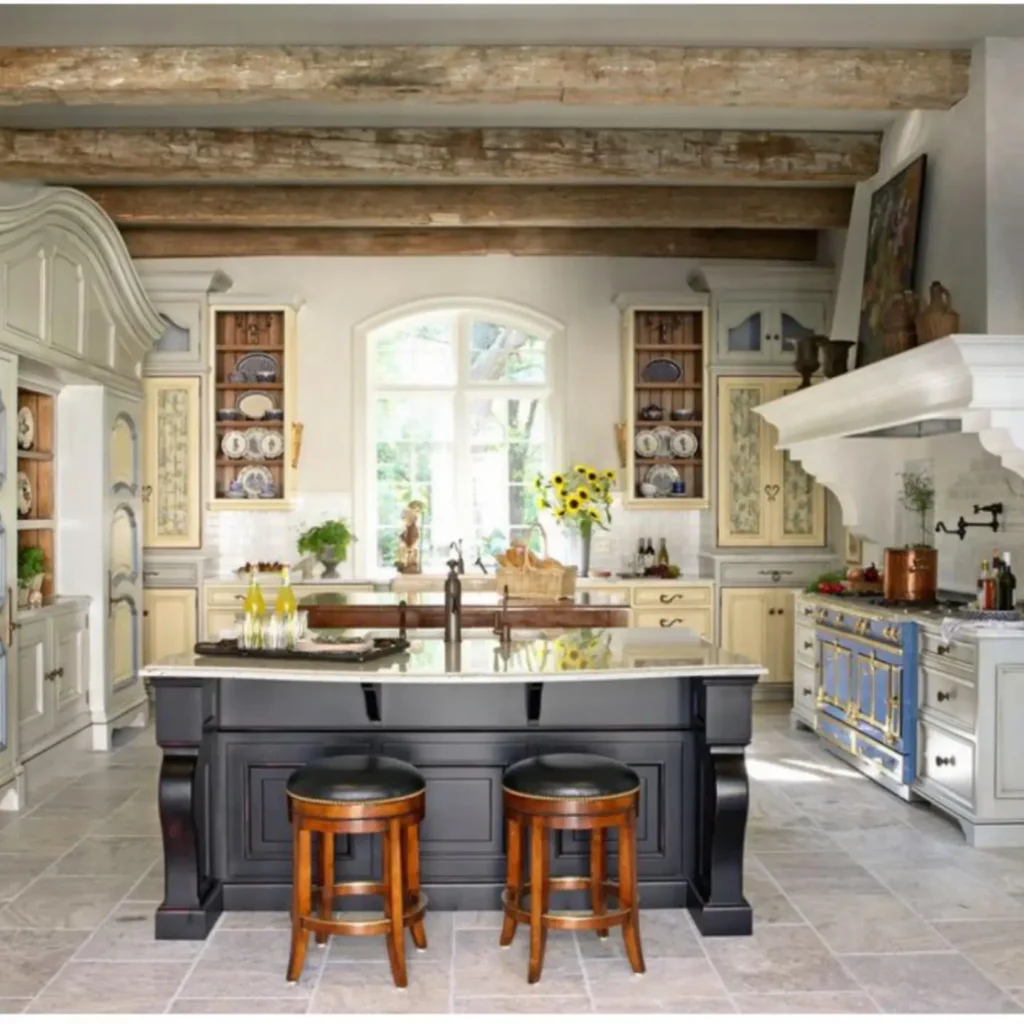 Spacious kitchen with exposed wooden ceiling beams, a marble-topped island with two stools, cream-colored cabinets, open shelves, and a large window letting in natural light, showcasing classic French country kitchen design elements.
