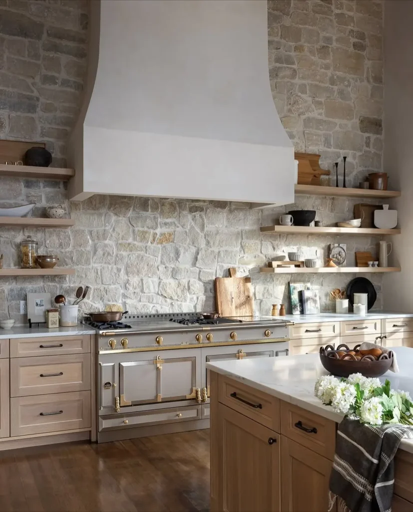 A modern french country kitchen features light wood cabinets, open shelves with dishes, a marble island, and a large stone backsplash with a white range hood above a modern stainless steel stove.