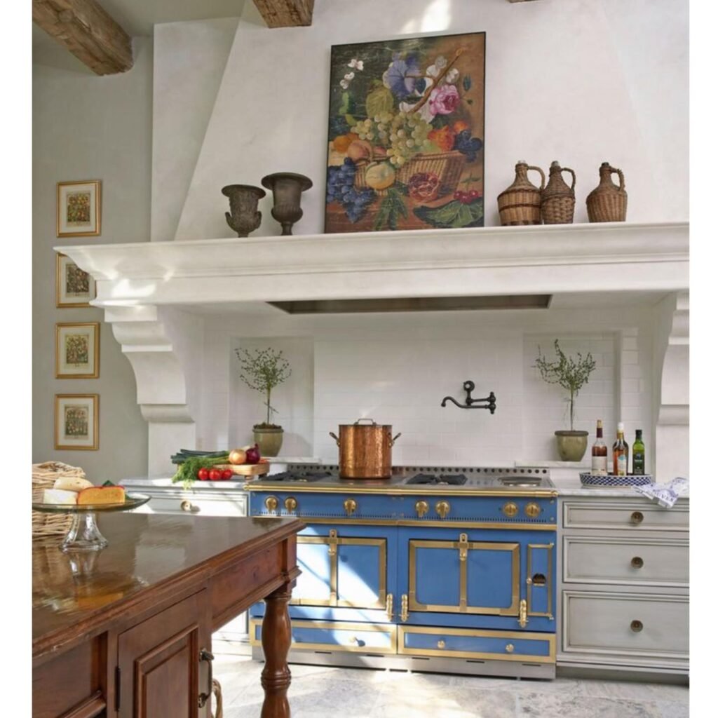 A french country kitchen with a blue and gold stove, copper pots, herbs, and baskets, featuring framed artwork and pottery displayed on a white tiled backsplash and mantle.
