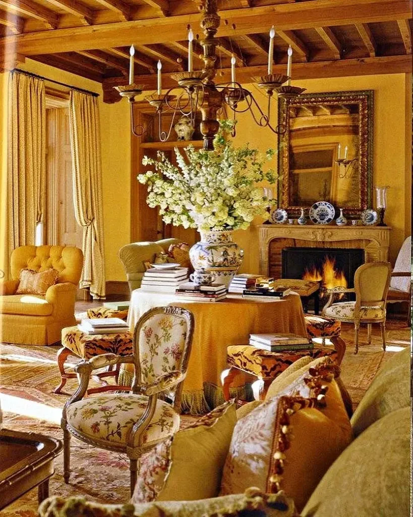 Elegant french country living room with gold tones, ornate chairs, a central round table with floral arrangement, fireplace, chandelier, and a large mirror above the mantel.