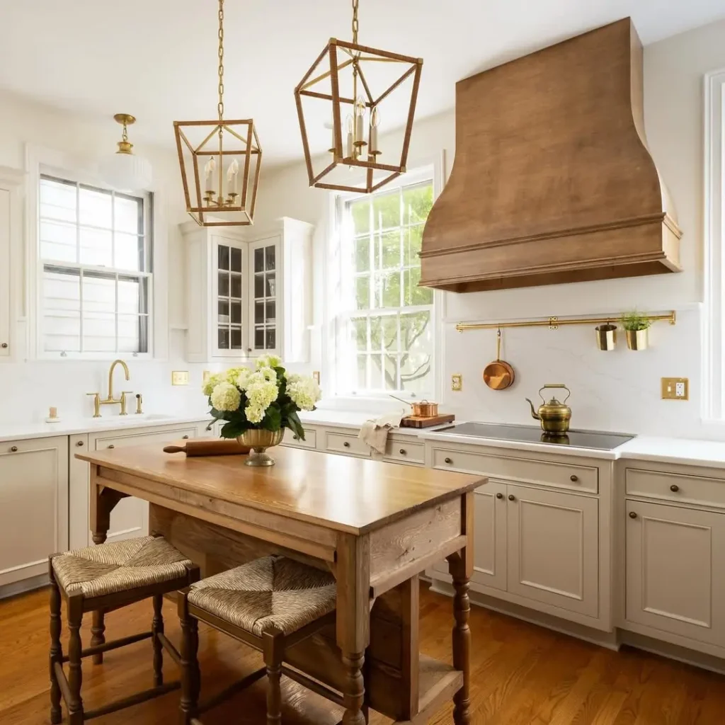 Bright french country kitchen with white cabinets, a wooden island, gold fixtures, large windows, and a rustic wooden range hood. A vase of white flowers sits on the island.