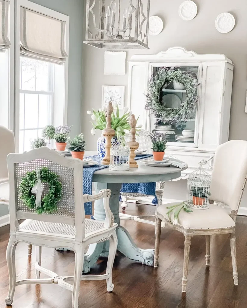 A light-filled dining area with a round table, white chairs, greenery, potted plants, and decorative wreaths, featuring rustic and vintage-style French country decor.