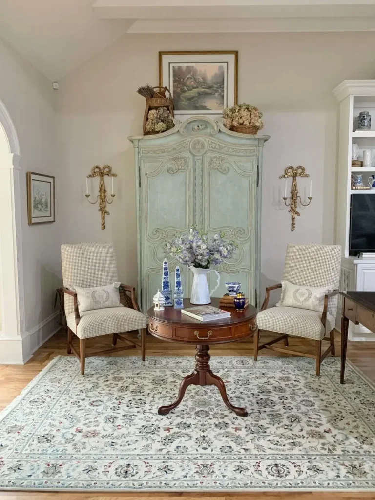 Elegant sitting area with two armchairs, a round wooden table, floral centerpiece, ornate armoire, decorative vases, and pastel colors on a patterned rug, perfectly designed to avoid common French Country decorating mistakes.