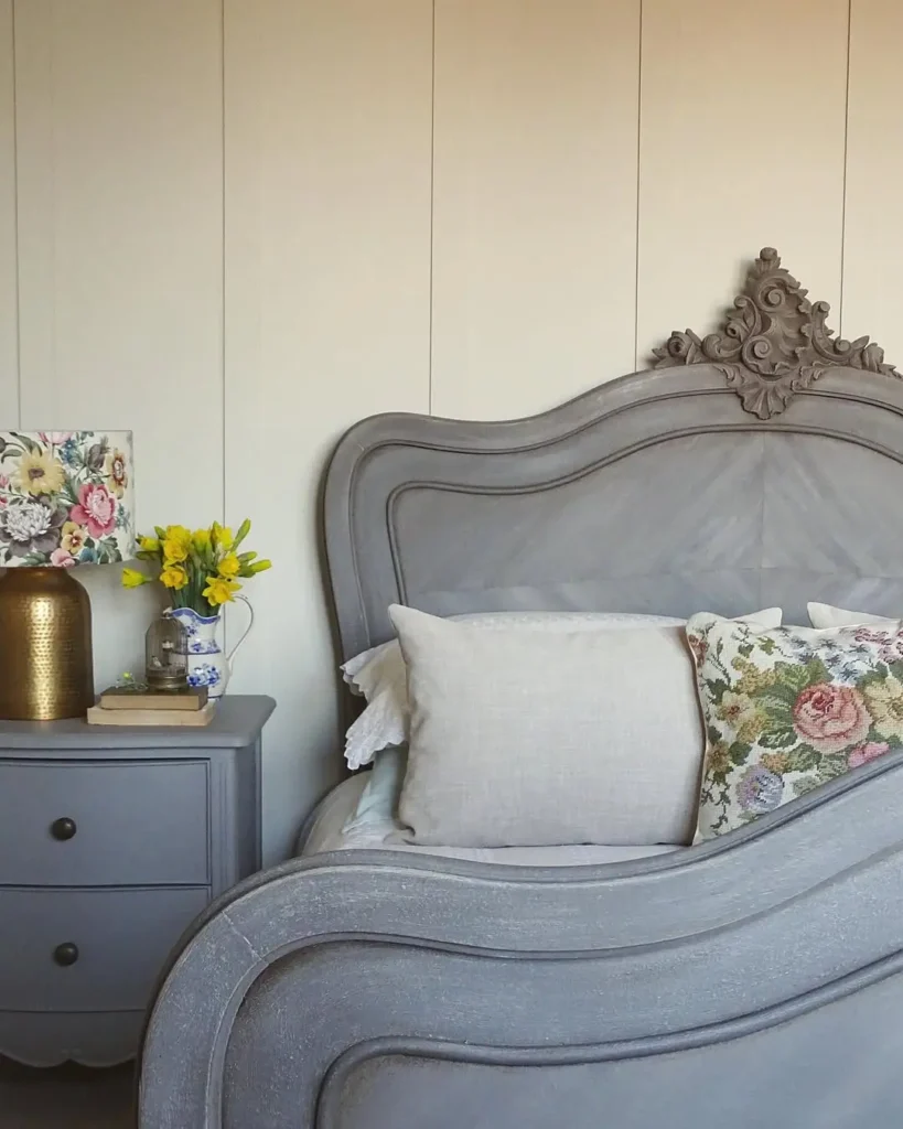 A slate vintage-style bed with white and floral pillows is beside a matching nightstand, set against a cream panel wall, a floral lamp, gold vase of yellow flowers, and books in this french country inspired bedroom.