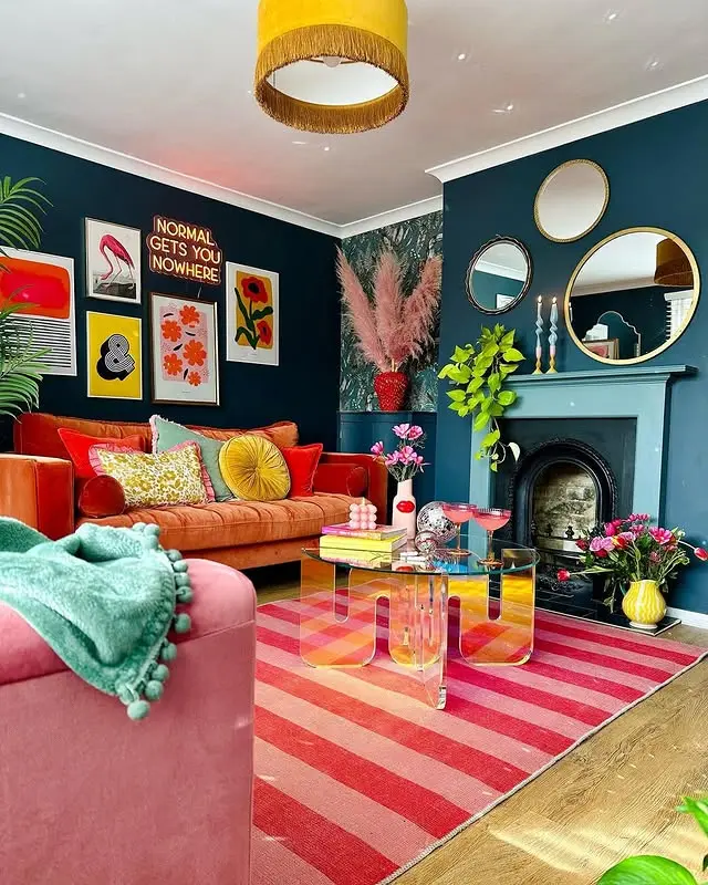 Colorful Funky Bohemian Living Room Decor with dark blue walls, an orange sofa, pink armchair, striped rug, wall art, plants, round mirrors, and a yellow ceiling light.