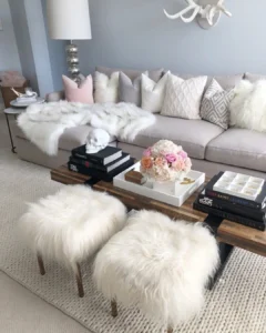 A modern living room styled for winter with a light gray sofa, white fur throws, fluffy stools, stacked books, a floral arrangement, and decorative accents on a coffee table.