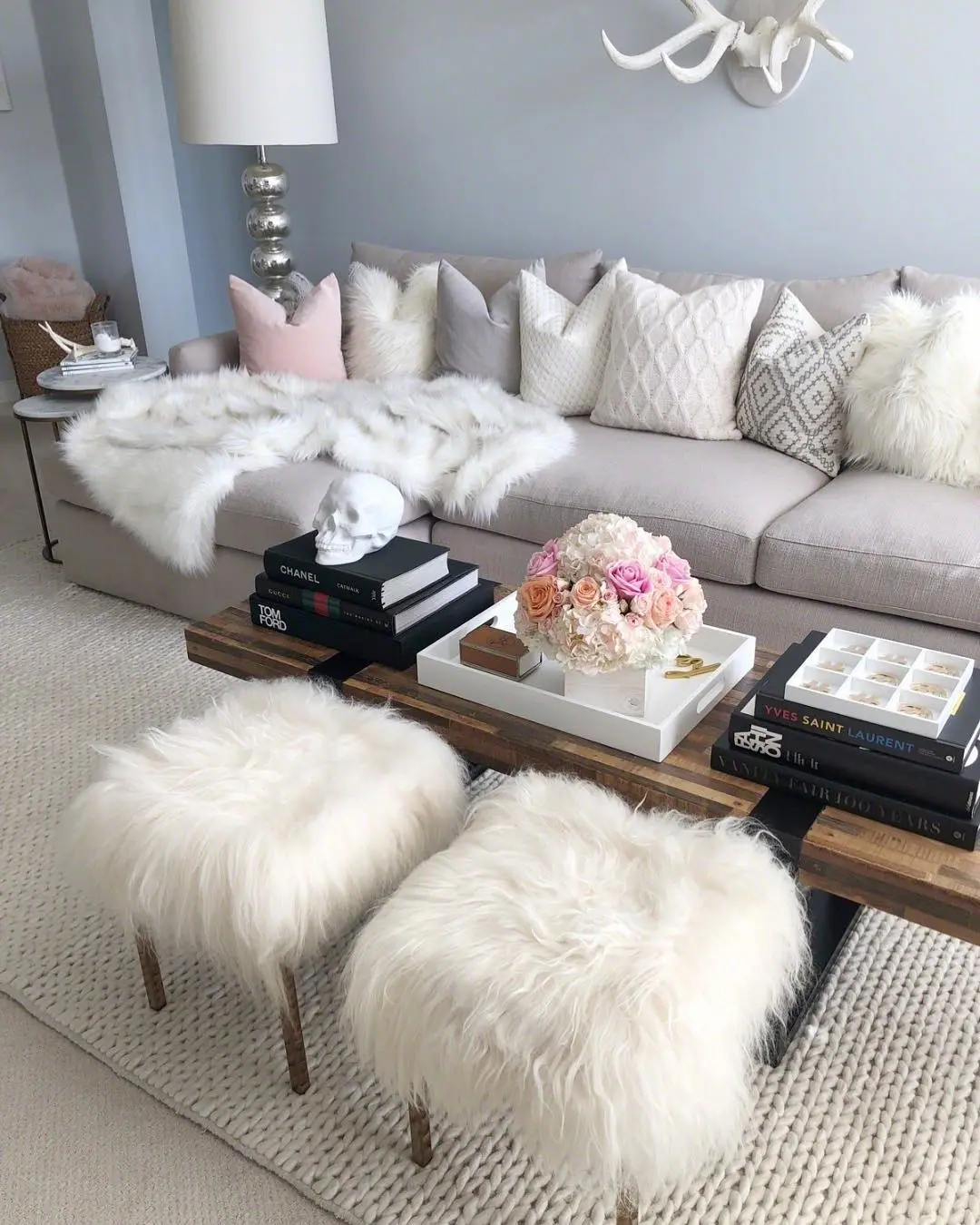 A modern living room styled for winter with a light gray sofa, white fur throws, fluffy stools, stacked books, a floral arrangement, and decorative accents on a coffee table.