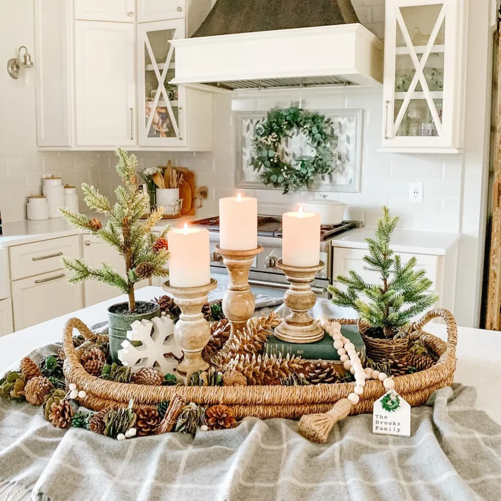 A woven basket with candles, small pine trees, pinecones, and garland creates a charming winter table vignette on the kitchen island. The kitchen features white cabinets and a wreath on the stove hood.