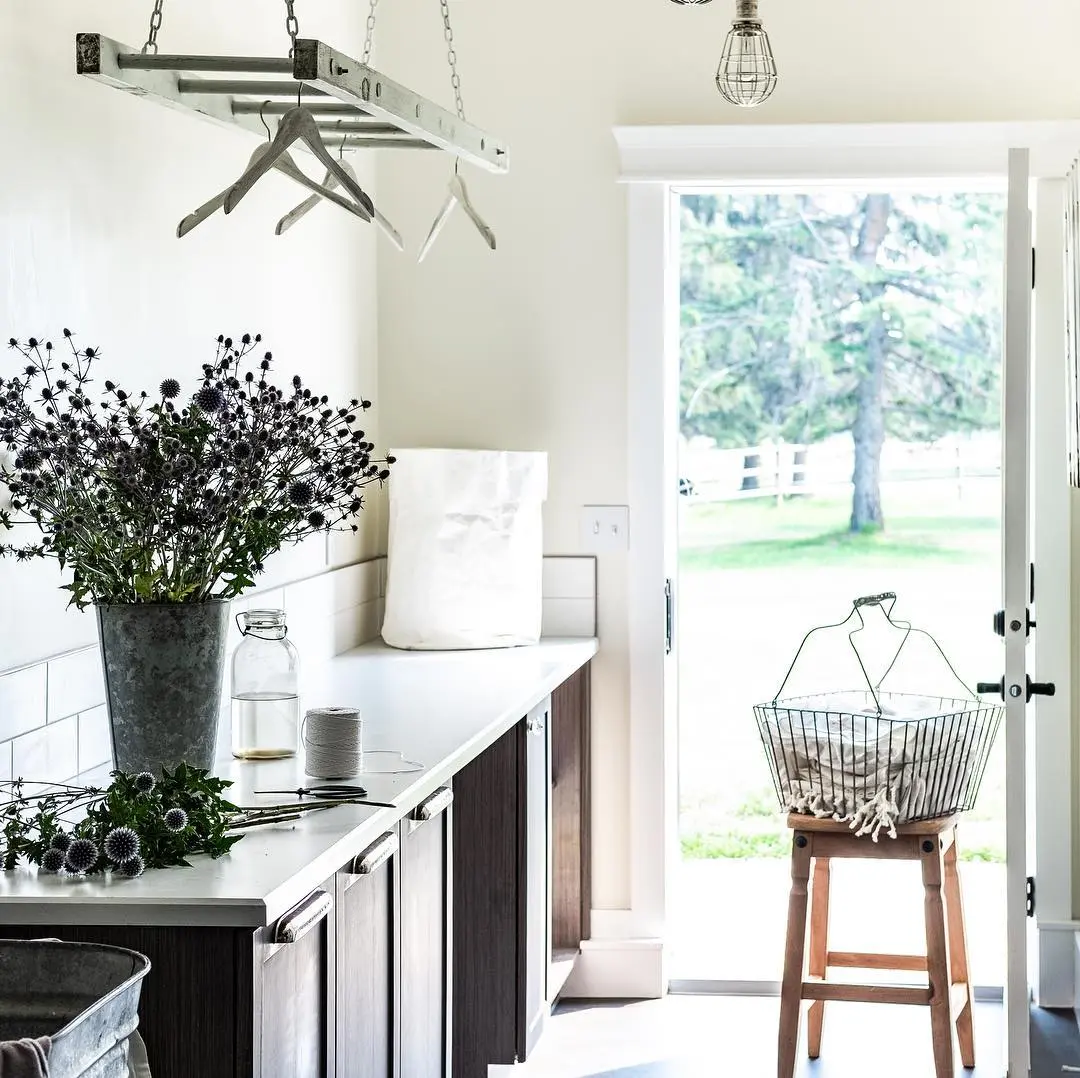 A bright laundry room with dark cabinets, a ladder propped by the counter to use as drying rack, a bucket of thistle flowers, a wire basket of towels on a wooden stool, and a door open to a green yard.