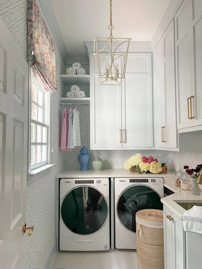 A laundry room with a washer and dryer, white cabinets, a hanging light fixture, folded towels, hanging clothes, and decorative flowers on the counter.