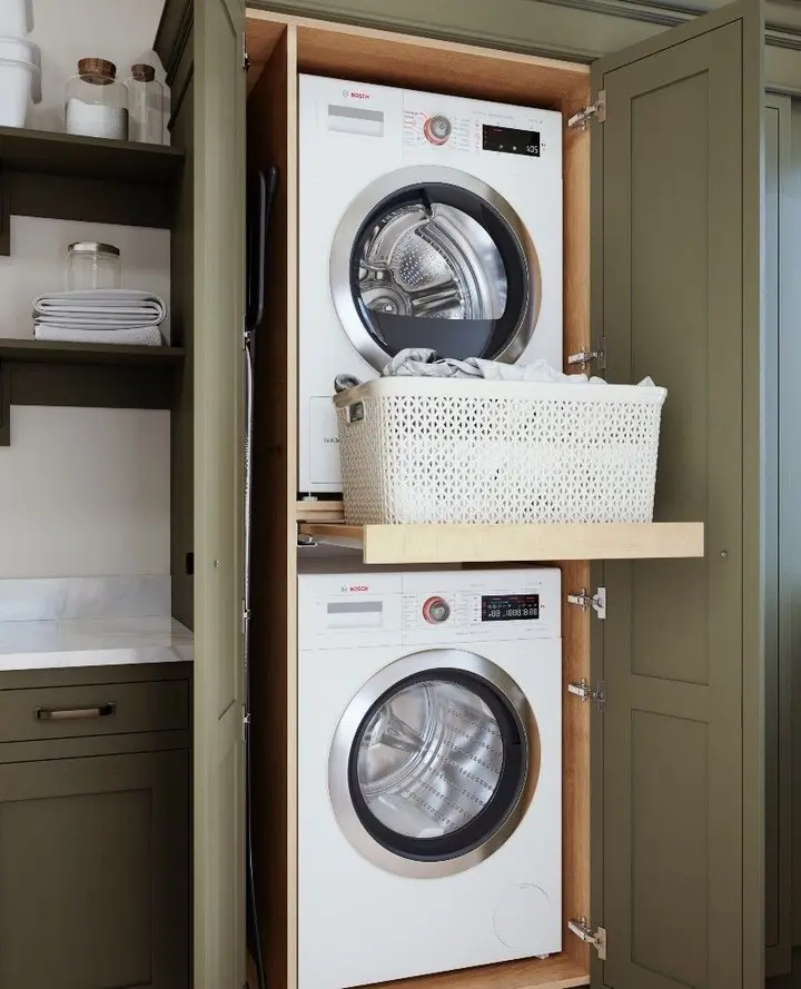 A stacked washer and dryer are enclosed in a cabinet with olive green doors. A pull-out folding shelf holds a white laundry basket between the two machines.