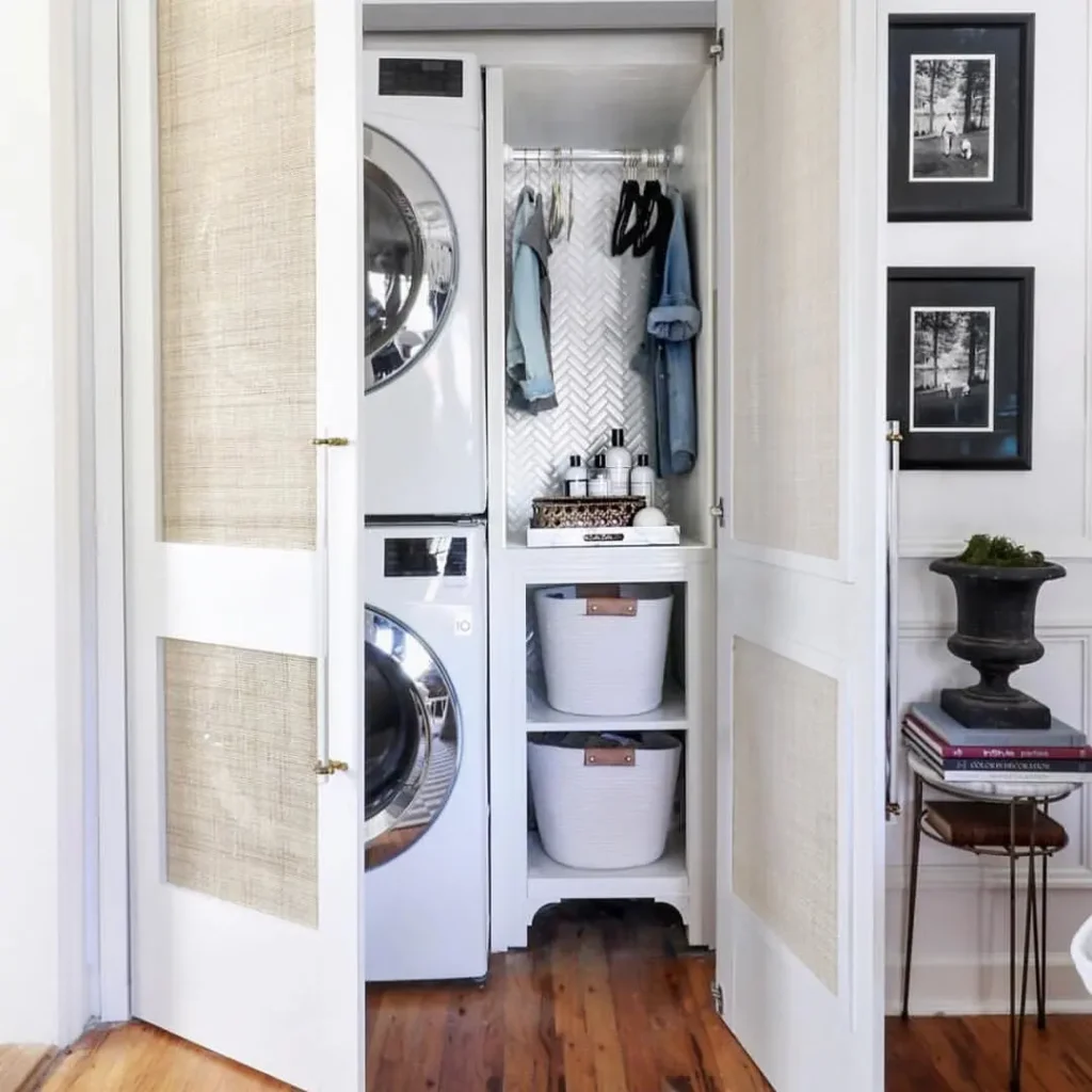 A compact laundry closet with a stacked washer and dryer, hanging clothes, two storage baskets, and cleaning supplies, partially concealed by open double doors.