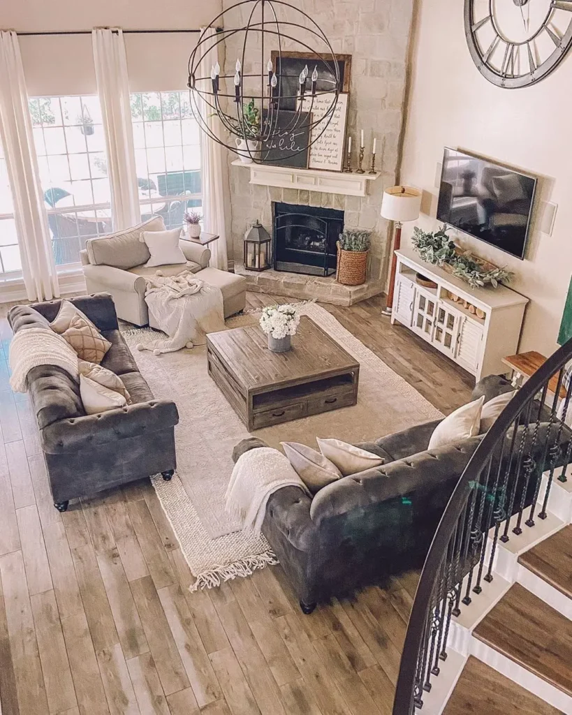 A spacious living room with gray sofas around layered rugs, a wooden coffee table, fireplace, wall-mounted TV, large windows, and a round chandelier, decorated in neutral tones.