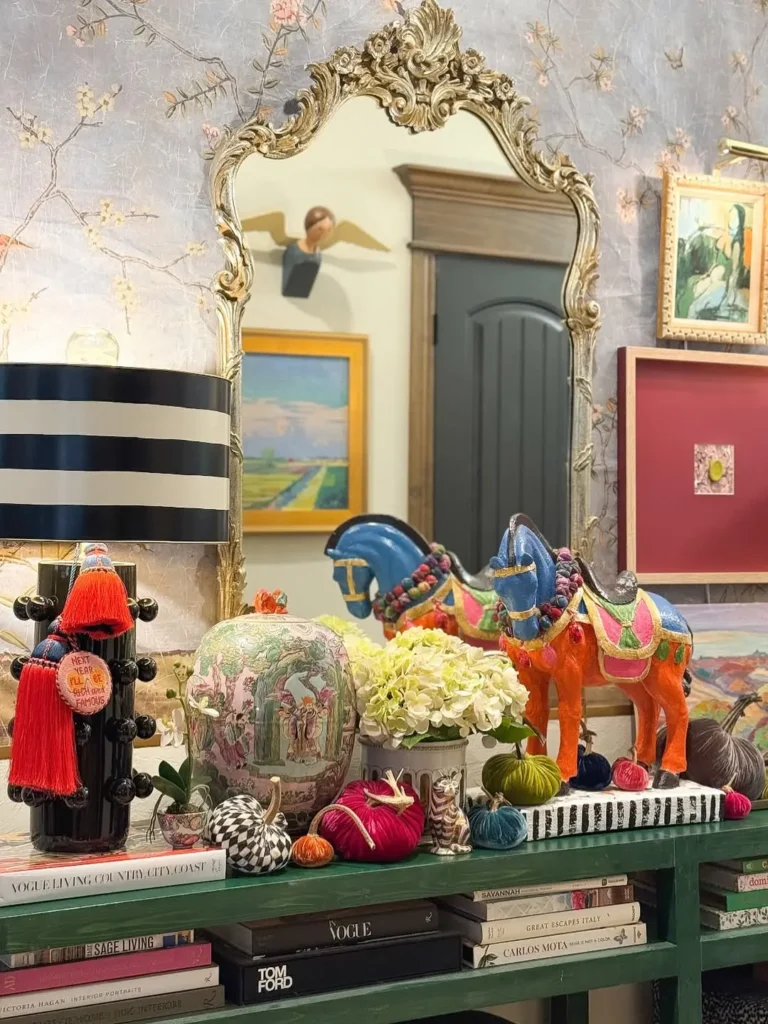 A maximalist fall entryway with an ornate mirror reflects colorful framed art above a shelf with books, decorative horses, yarn, flowers, and various eclectic ornaments arranged together.