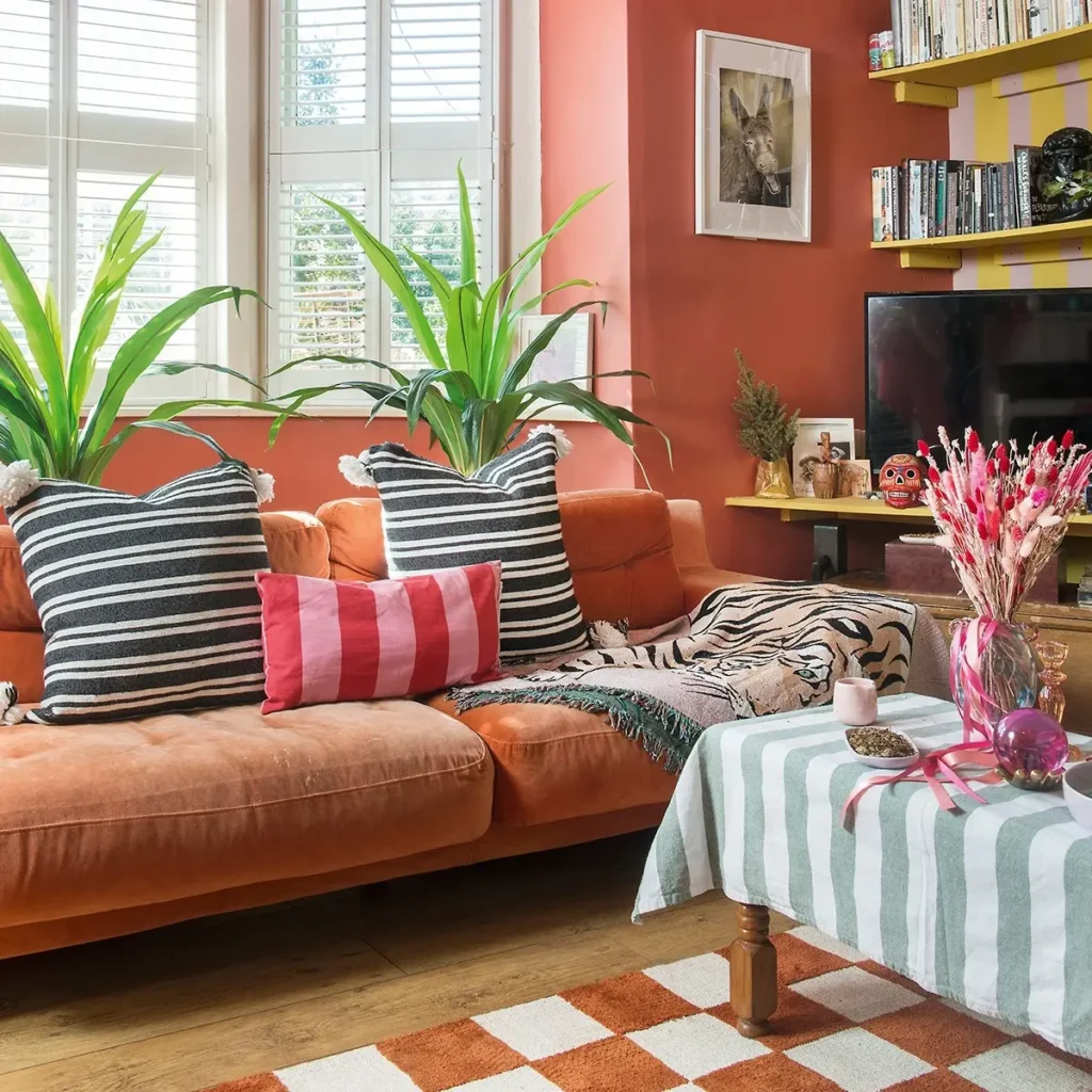 A maximalist living room with an orange sofa, striped cushions, potted plants, a coffee table with a striped cloth, and shelves filled with books and decor against a bold pink wall.