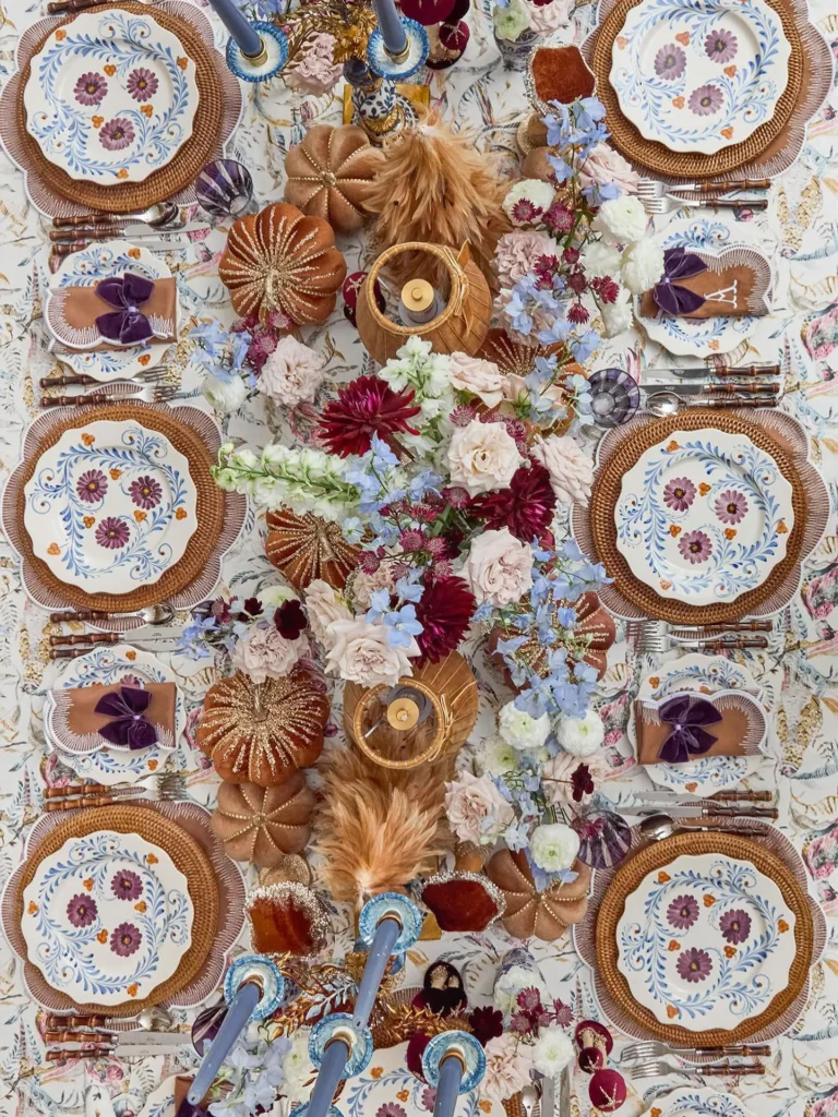 A maximalist fall tablescape features a decorated dining table set for six with floral plates, woven placemats, blue candles, small decorative pumpkins, and an arrangement of flowers in rich autumn colors.