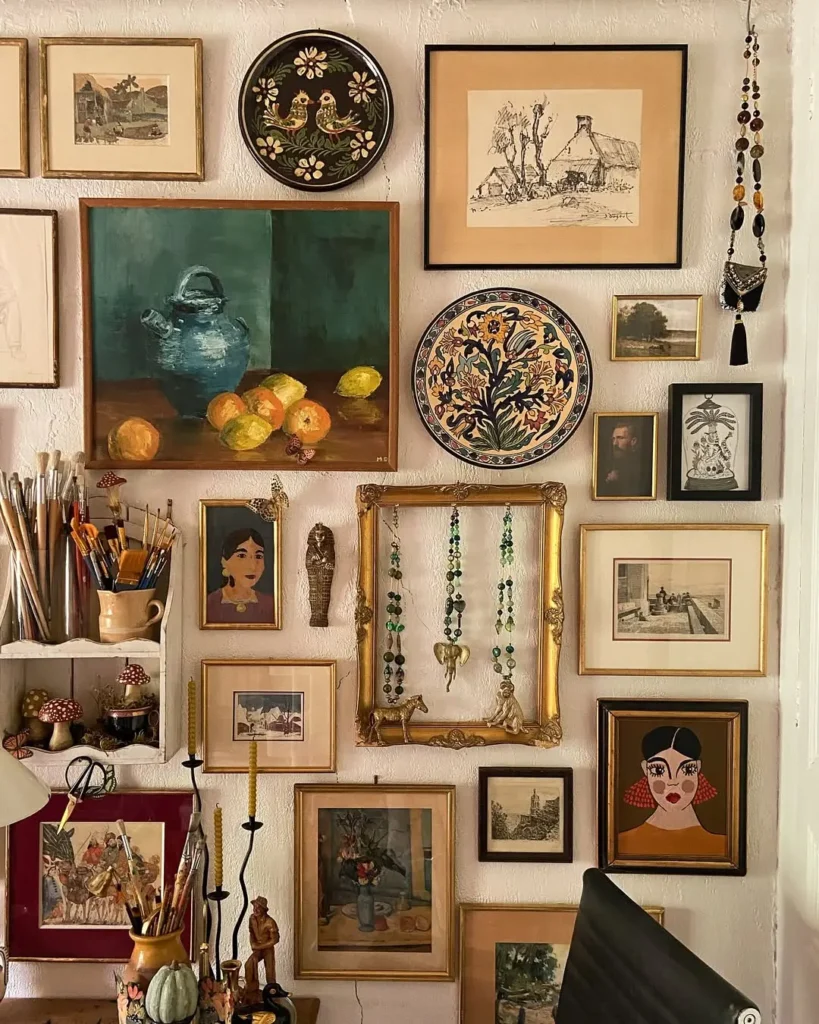 A wall filled with assorted framed artwork, paintings, decorative plates, necklaces, and paintbrushes showcases the art of choosing and placing wall art in a maximalist home within a cozy, creative workspace.