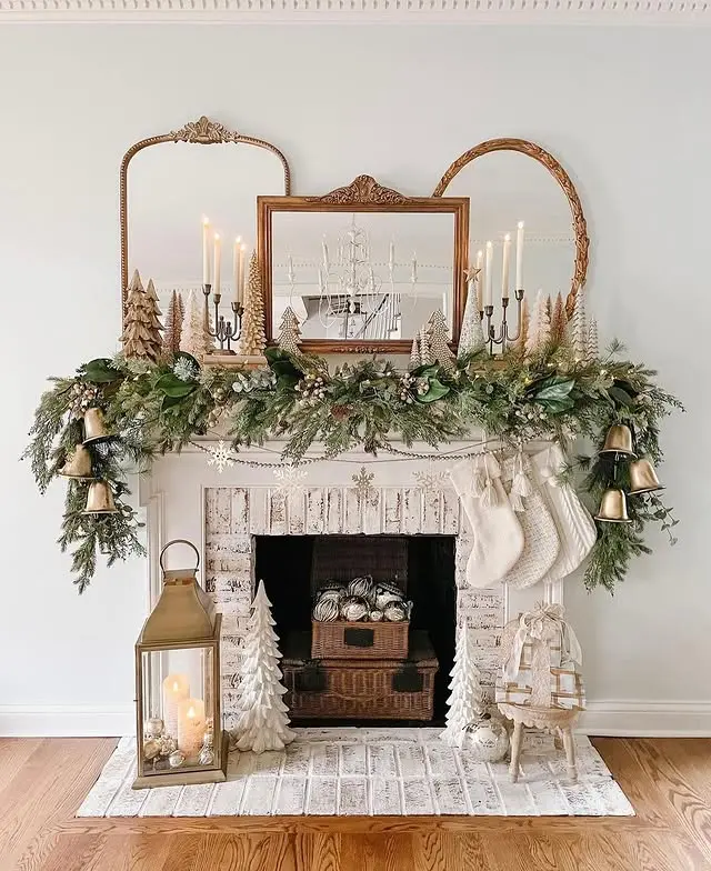 A decorated white fireplace with greenery, candles, stockings, gold bells, mirrors, and ornaments, featuring a lantern with candles and small decorative trees on the hearth.