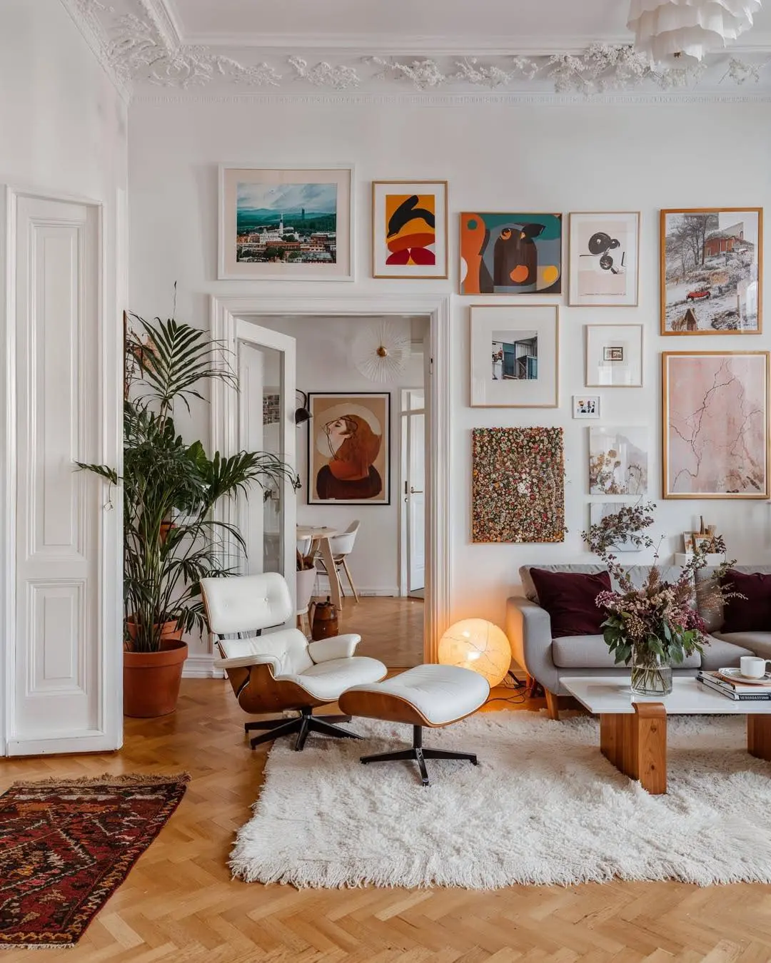 A mid-century modern-inspired living room featuring an iconic Eames lounge chair with ottoman on a shag rug, a sofa, indoor plants, a wooden coffee table, and a gallery wall of colorful art and photos above the sofa.