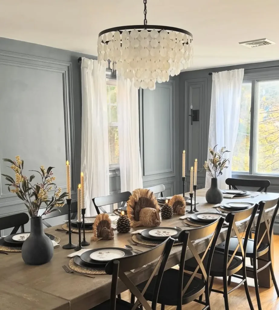 A dining room with a wooden table set for eight, black chairs, decorative vases, pinecones, and candles under a white chandelier creates a minimalist Thanksgiving setting filled with natural light from two large windows.