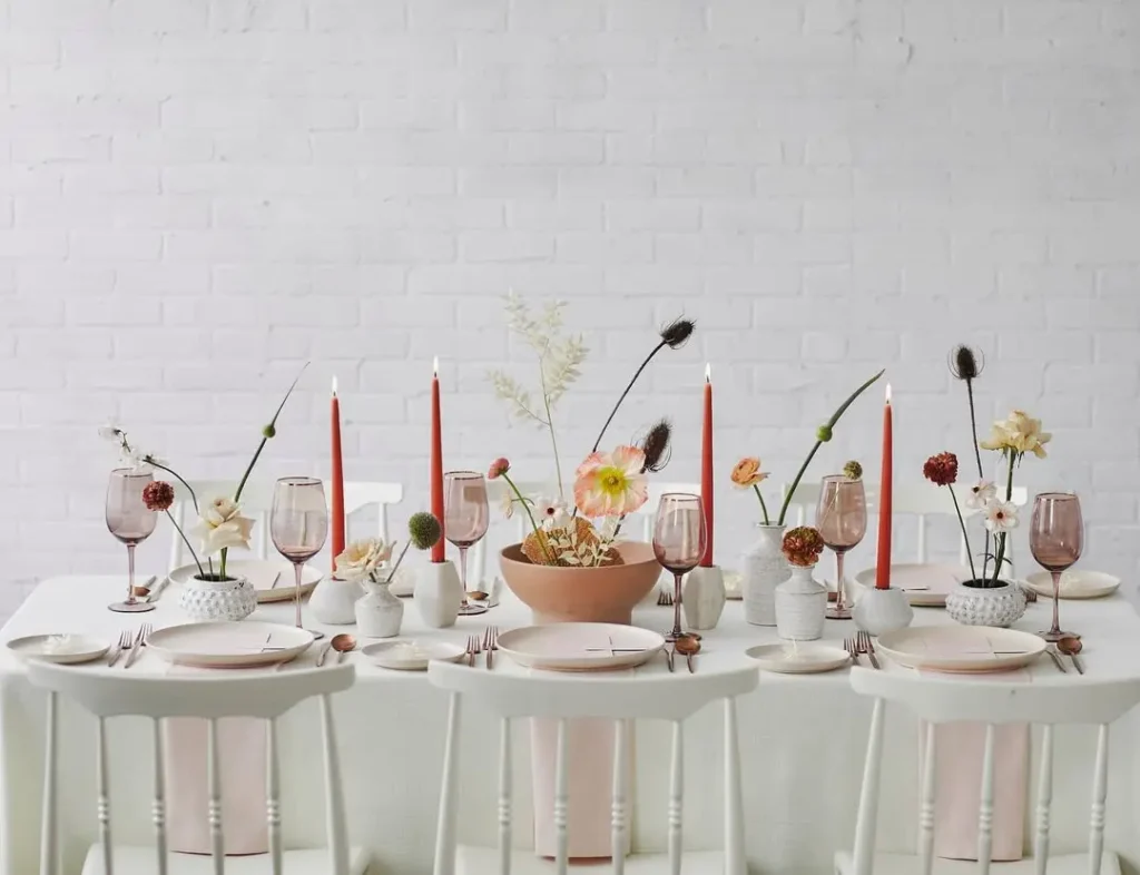 A neatly set dining table with white chairs, pink-tinted glassware, red candles, and minimalist floral centerpieces creates a perfect setting for a minimalist Thanksgiving against a white brick wall.