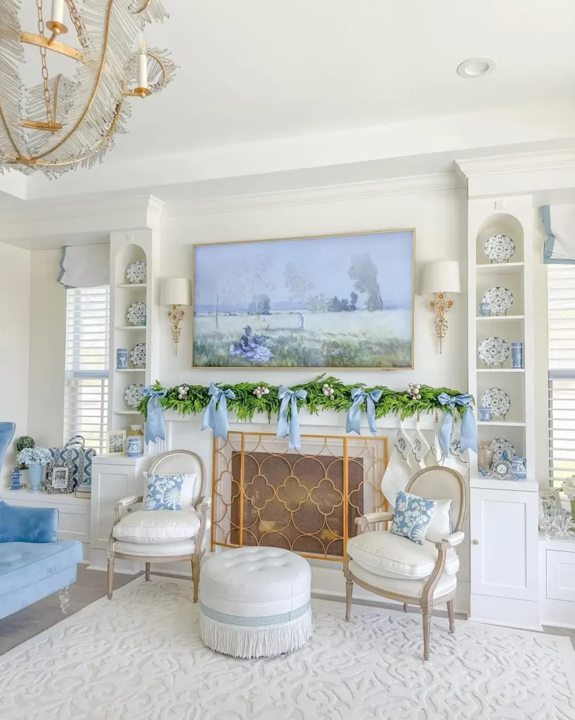 Elegant living room with a misty blue & white French country color palette, two armchairs, a round ottoman, a fireplace adorned with greenery and blue ribbons, and shelves with decorative items on either side.