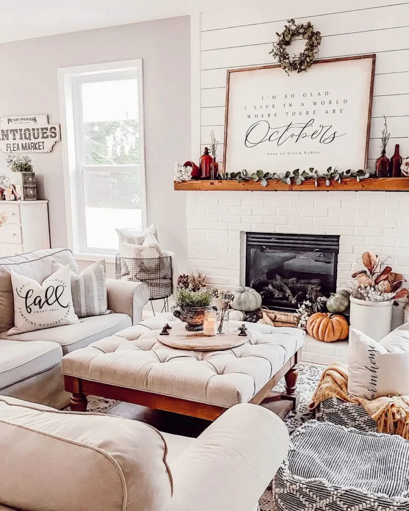 A cozy farmhouse living room with neutral furniture, fall-themed decor, pumpkins, a mantel with a wreath, and a large framed quote above a white brick fireplace.