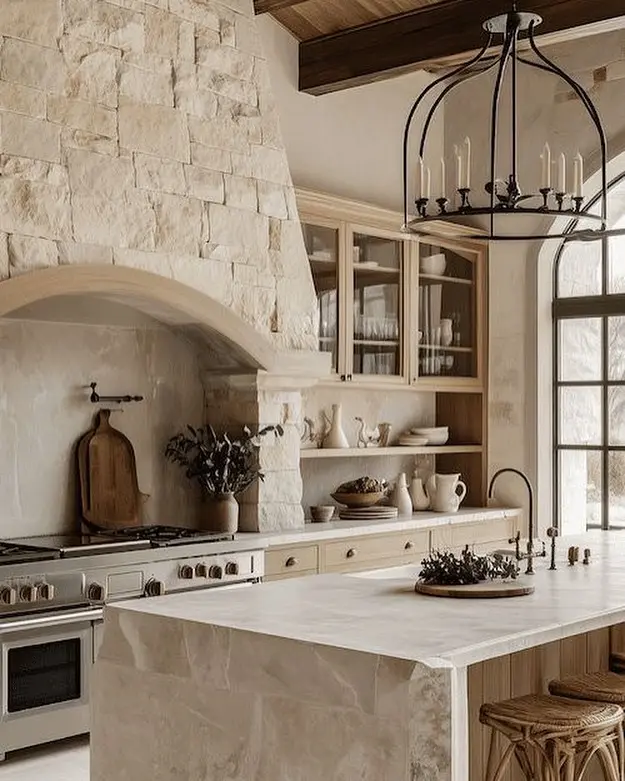 A rustic French Country Kitchen with modern appliances, featuring stone walls, wooden cabinets, open shelves, a large island with stools, and a black metal chandelier.