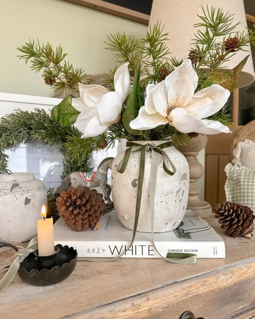 A rustic vase with white flowers and greenery sits on a book titled "White," next to pinecones, a small lit candle, and other winter decor on a wooden surface.